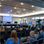 A group of people sit in chairs in a meeting room. Others are sitting at the head table. The screen says, "Grand Valley State University Board of Trustees meeting, February 21, 2025.